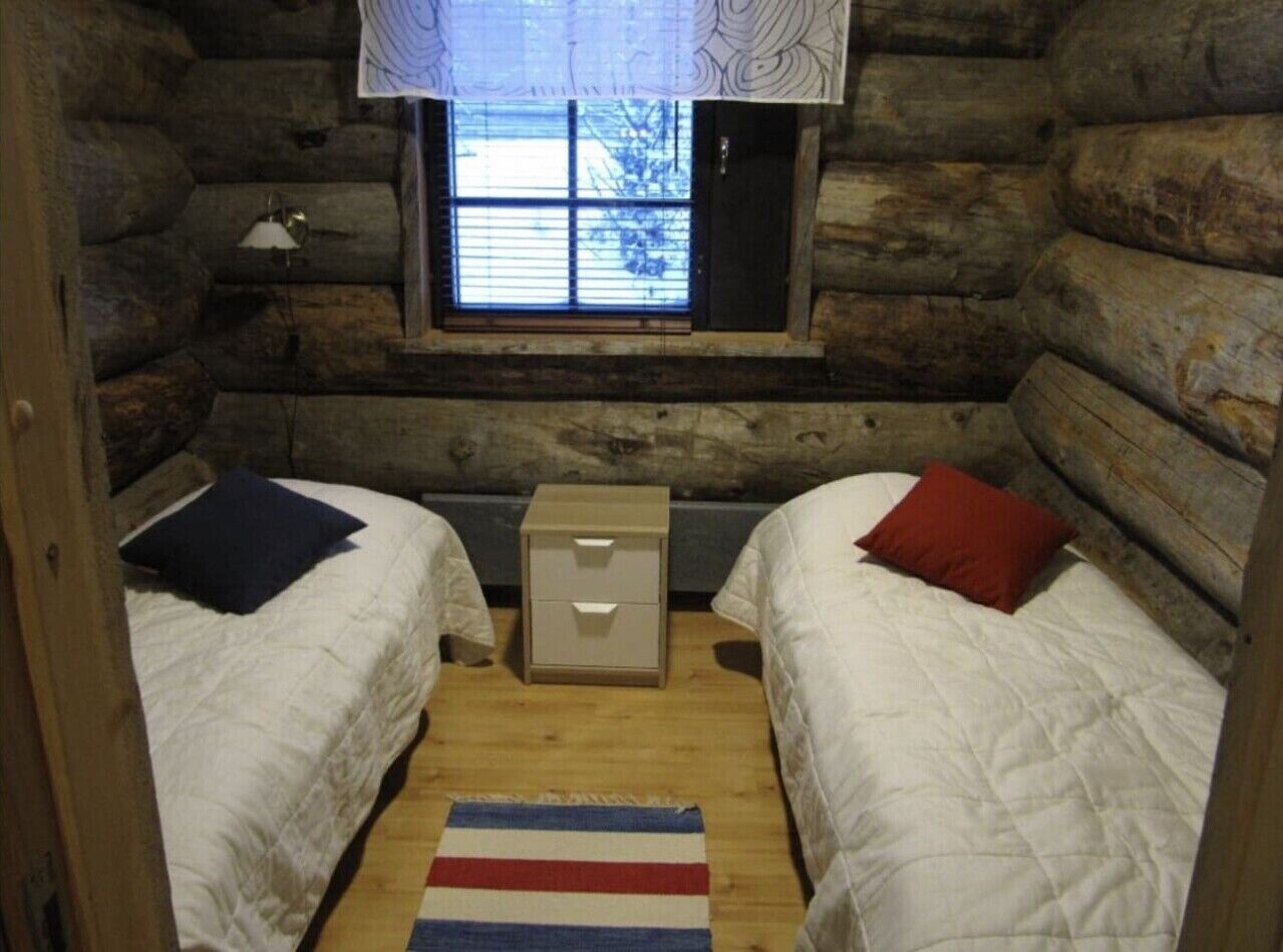 A log cabin bedroom with two beds, a window, a small bedside table, and a striped rug.