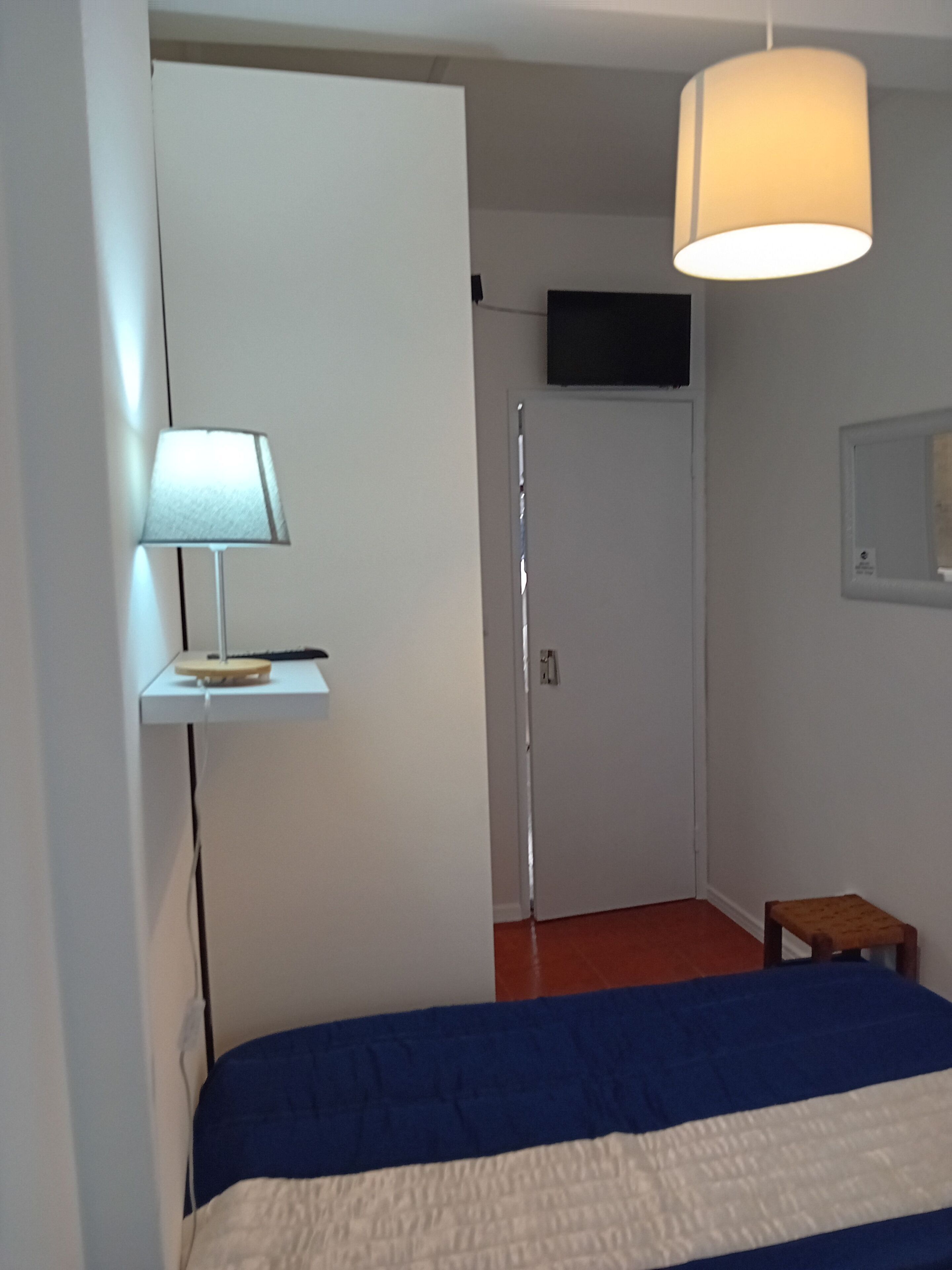 Une chambre à coucher avec un lit, une lampe de chevet, un téléviseur fixé au mur et un petit tabouret en bois.