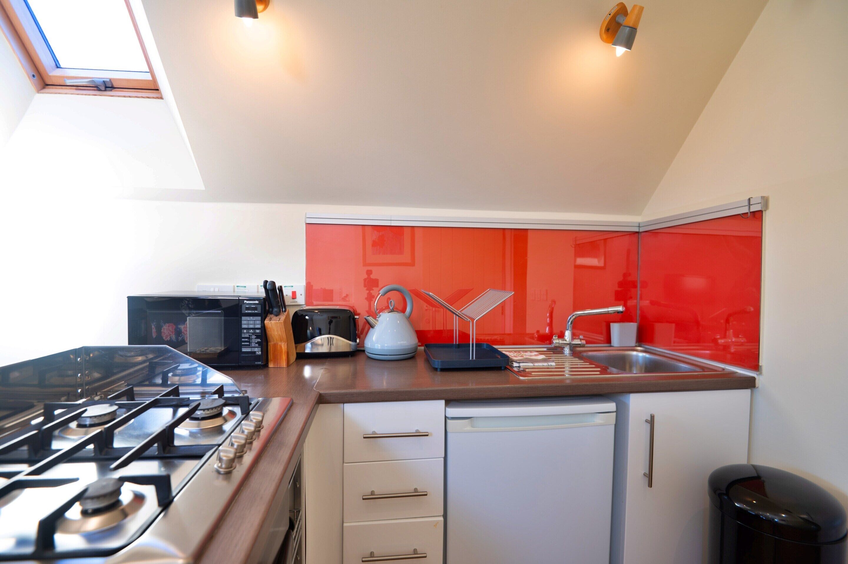 A compact kitchen with a gas stove, a microwave, a toaster, and a kettle. There is a sink with a faucet, and a dishwasher is visible. The kitchen has a red backsplash and white cabinets.