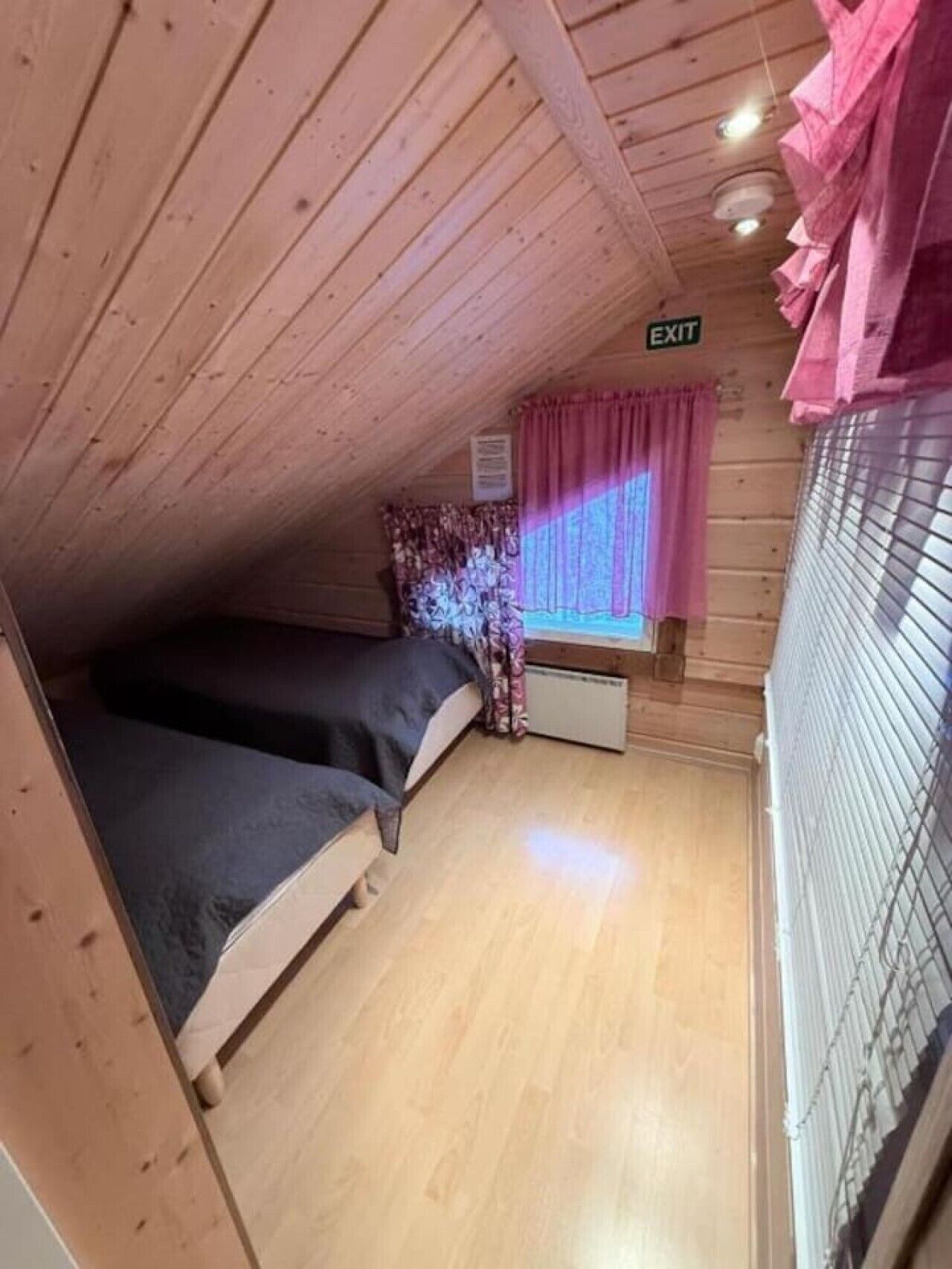 A small, enclosed room with a bunk bed, a window with curtains, an exit sign, and a radiator.
