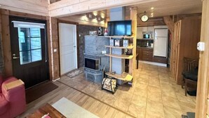 A spacious living area with a fireplace, wooden shelves, and a kitchen in the background. - Rukasoihtu A | 2BR Cabin with Sauna & Fireplace (Kuusamo)