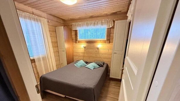 A bedroom with a bed, curtains, a window, and a wall-mounted light fixture. - Rukasoihtu A | 2BR Cabin with Sauna & Fireplace (Kuusamo)