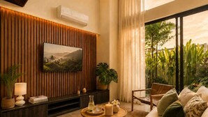 A modern living room with a large flat-screen TV, a wooden accent wall, a cozy seating area with a sofa and armchairs, a round coffee table, and a view of lush greenery through large windows. - Villa Gaudi II (Kabupaten Badung)