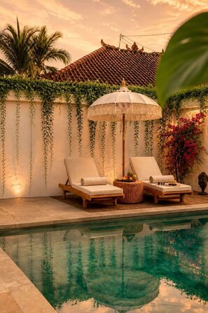 A poolside area with two lounge chairs, a decorative umbrella, and a wall adorned with hanging plants and flowers. - Villa Juma (Kabupaten Badung)