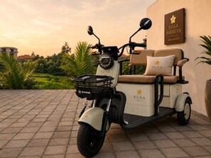 A stationary electric scooter with a seat and a basket, parked on a paved surface with a building in the background. - Villa Hibiscus I (Kabupaten Badung)