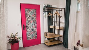 Um quarto com uma porta rosa, uma planta, uma estante de madeira e piso branco.