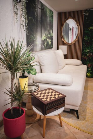 Una sala de estar moderna con un sofá blanco, una pequeña mesita de madera con un tablero de ajedrez y una planta en maceta.