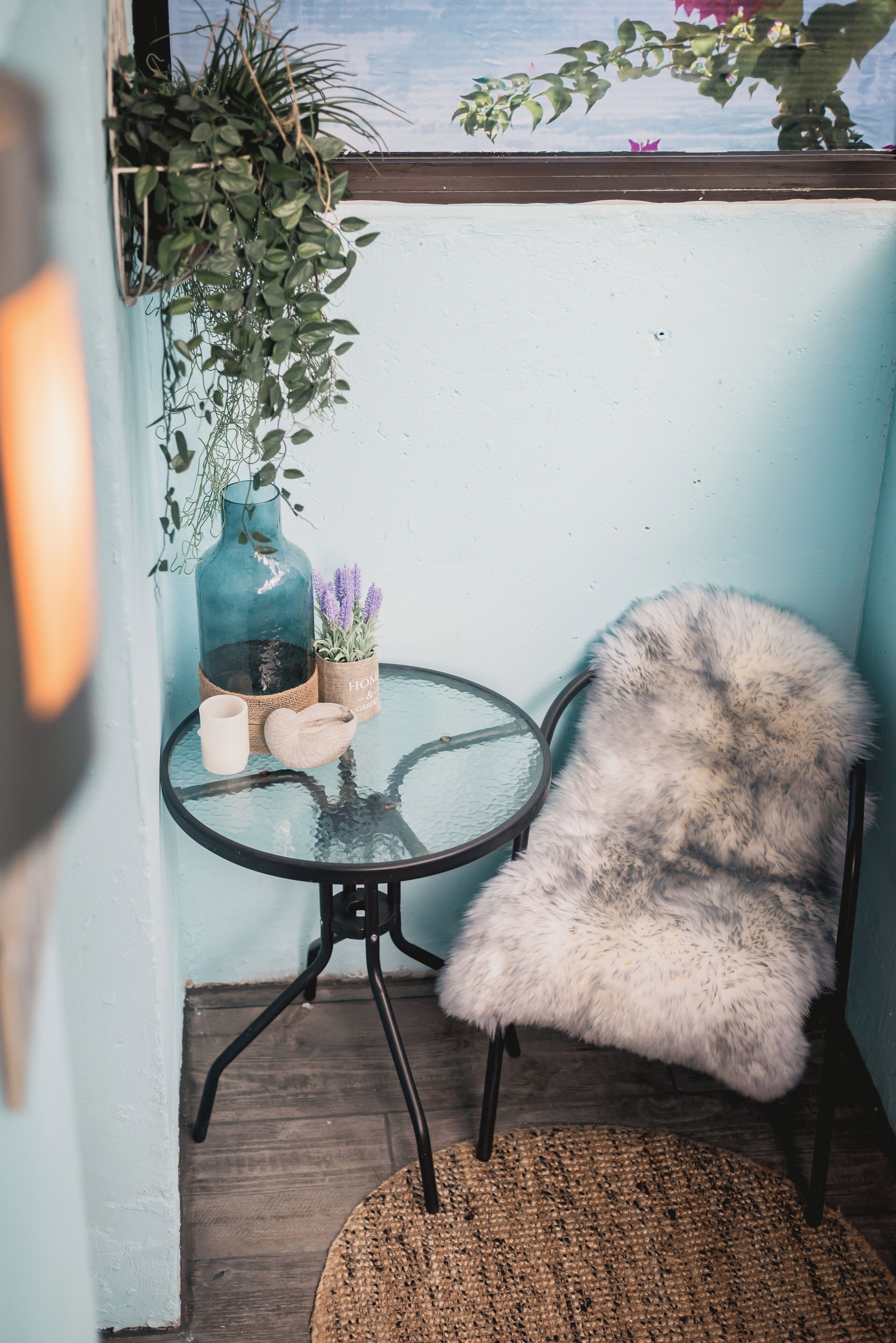 Ein kleiner runder Tisch mit Glasplatte, ein flauschiger weißer Sessel, ein gewebter Teppich, eine blaue Vase mit lila Blumen und eine hängende Pflanze.