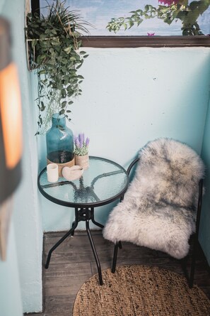 Ein kleiner runder Tisch mit Glasplatte, ein flauschiger weißer Sessel, ein gewebter Teppich, eine blaue Vase mit lila Blumen und eine hängende Pflanze.