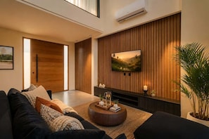 A modern living room with a large flat-screen TV, a dark-colored sofa, a round coffee table with decorative items, and a wooden accent wall. - Villa Gaudi I (Kabupaten Badung)