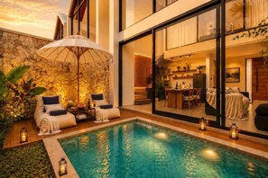 A modern pool area with a stone wall, a lounge chair, and a dining area visible through a glass door. - Villa Gaudi I (Kabupaten Badung)