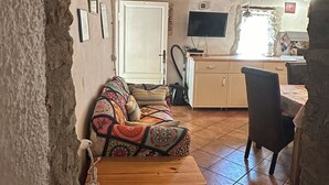Un interior rústico con un sofá, un cofre de madera, suelo de baldosas y una zona de cocina con televisión.