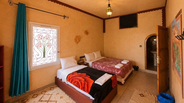 A bedroom with two beds, a window with a decorative design, a ceiling light, and a door leading to a bathroom. - Auberge Restaurant Zahra (Ait Sedrate Jbel El Soufla)