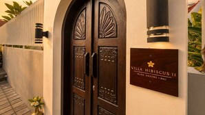 A wooden double door with intricate carvings, a wall-mounted sign, and a vertical light fixture. - Villa Hibiscus II (Kabupaten Badung)