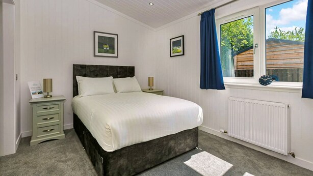 A bedroom with a bed, bedside tables, a window with curtains, and a radiator. - Lochside Perthshire Lodge for 4 (Perth)