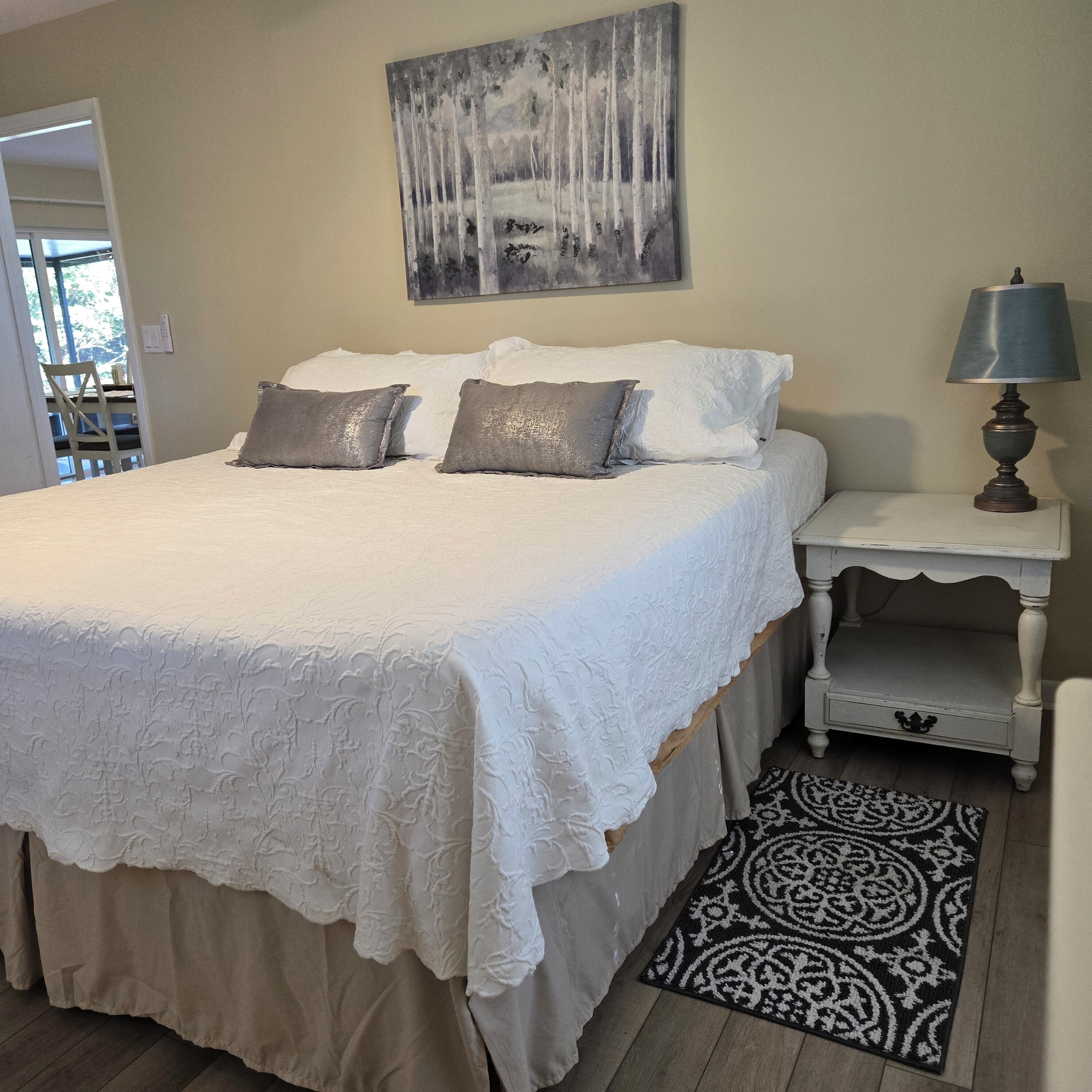 A bedroom with a bed, pillows, a bedside table with a lamp, a rug, and a painting on the wall.