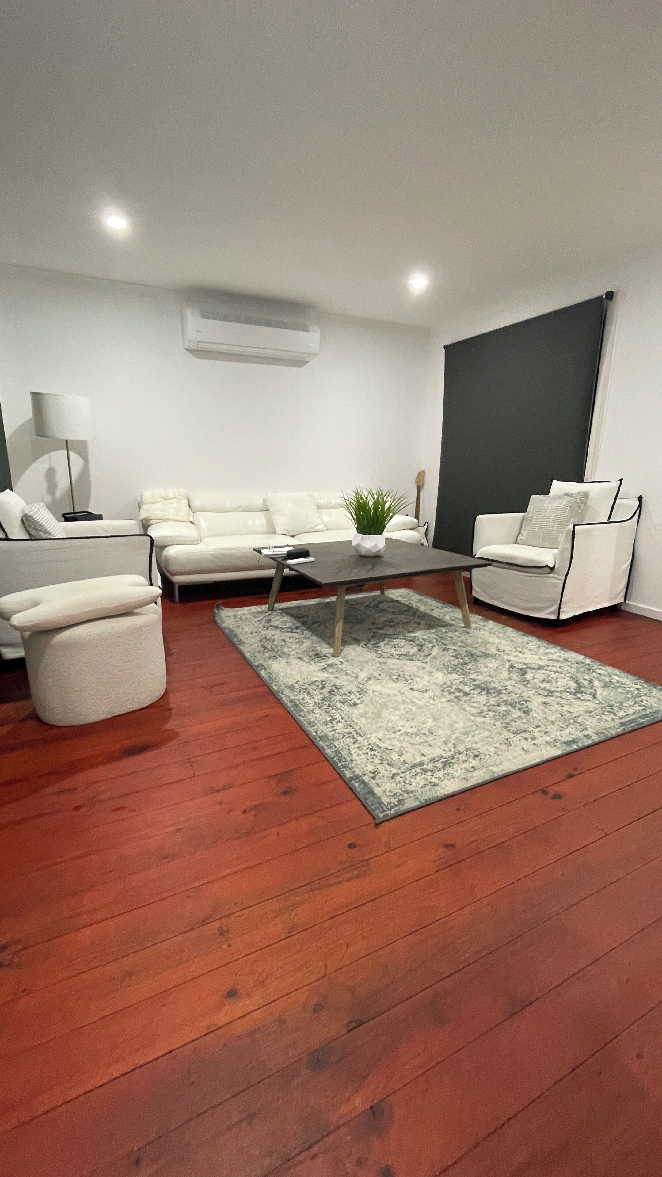 A modern living room with a white sofa set, a coffee table with a plant, and a large dark screen on the wall.