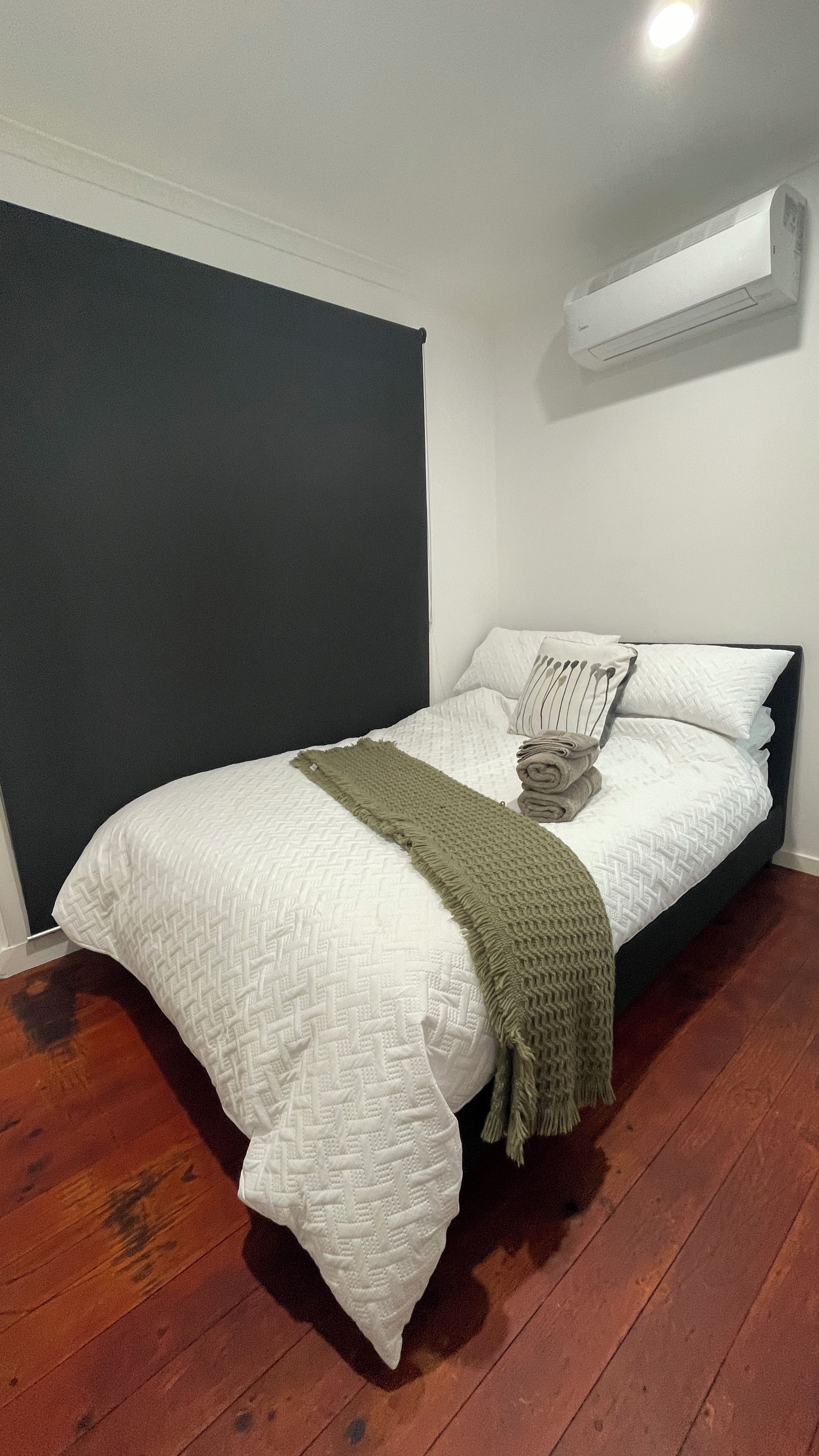 A bedroom with a bed, a dark accent wall, a white air conditioning unit, and a wooden floor.