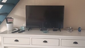 A white entertainment unit with a flat-screen TV, remote controls, and a cup with magazines. - Beautiful 2-Bedroom Townhouse, Frontignan, Secure Residential Complex, Beach Access 100 m Away (Frontignan)