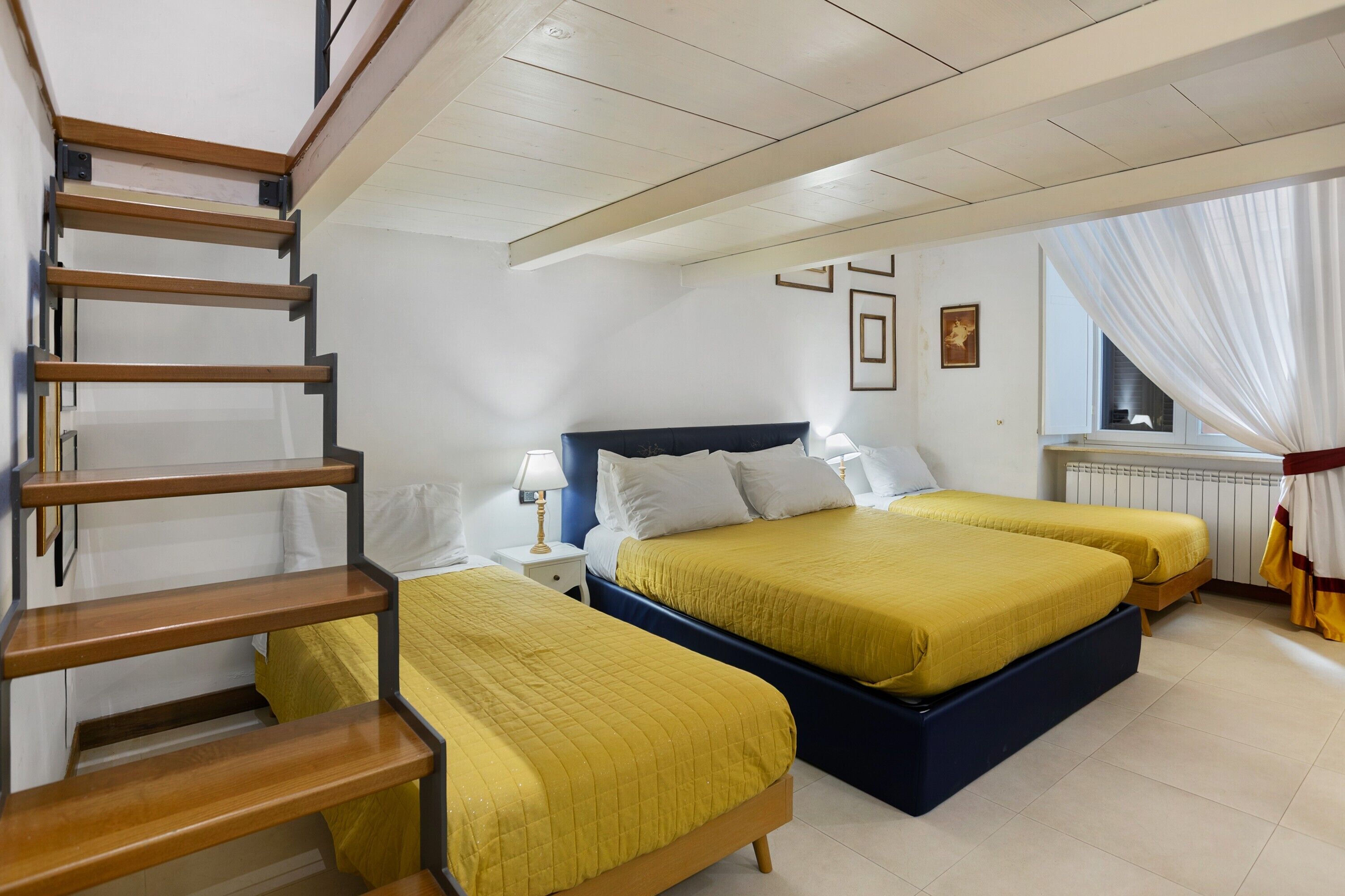 Een hotelkamer met een trap, twee bedden met gele beddengoed, een nachtkastje met een lamp en een raam met gordijnen.