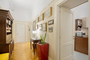 A hallway with a wooden floor, a bookshelf, a table with a lamp, a potted plant, and a kitchen area with cabinets and a microwave. - La Dimora Dei Reali – Yellow Suite with Wi-Fi and Air Conditioning (Naples)