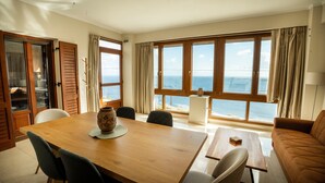 Une pièce comprenant une table à manger en bois, des chaises, un canapé et de grandes fenêtres donnant sur la mer.