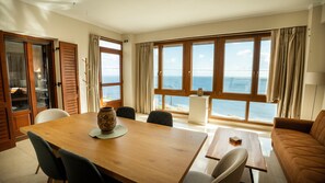 A room with a wooden dining table, chairs, a sofa, and large windows with a view of the sea. - Villa 'Vignetta Mare 2br Seaview Retreat' with Sea View, Wi-Fi and Air Conditioning (Ermoupoli)