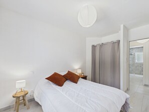 A bedroom with a bed, a bedside table with a lamp, a curtain, and a view into another room.