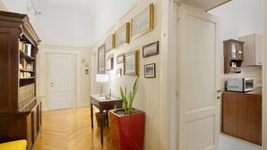 Een hal met een houten vloer, een boekenkast, een tafel met een lamp, een potplant, en een keuken met kastjes en een magnetron.