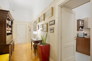 A hallway with a wooden floor, a bookshelf, a table with a lamp, a potted plant, and a kitchen area with cabinets and a microwave. - La Dimora Dei Reali - Bordeaux Suite with Private Terrace (Naples)