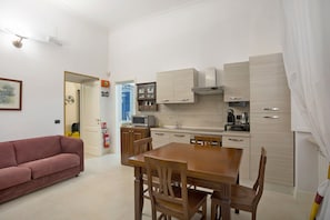 A compact living space with a kitchenette, a dining area with a wooden table and chairs, a sofa, and a door leading to a bathroom with a shower. - La Dimora Dei Reali - Bordeaux Suite with Private Terrace (Naples)