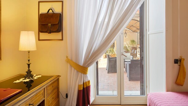 Een kamer met een bed, een bureau met lamp, een raam met gordijnen en uitzicht op een terras.