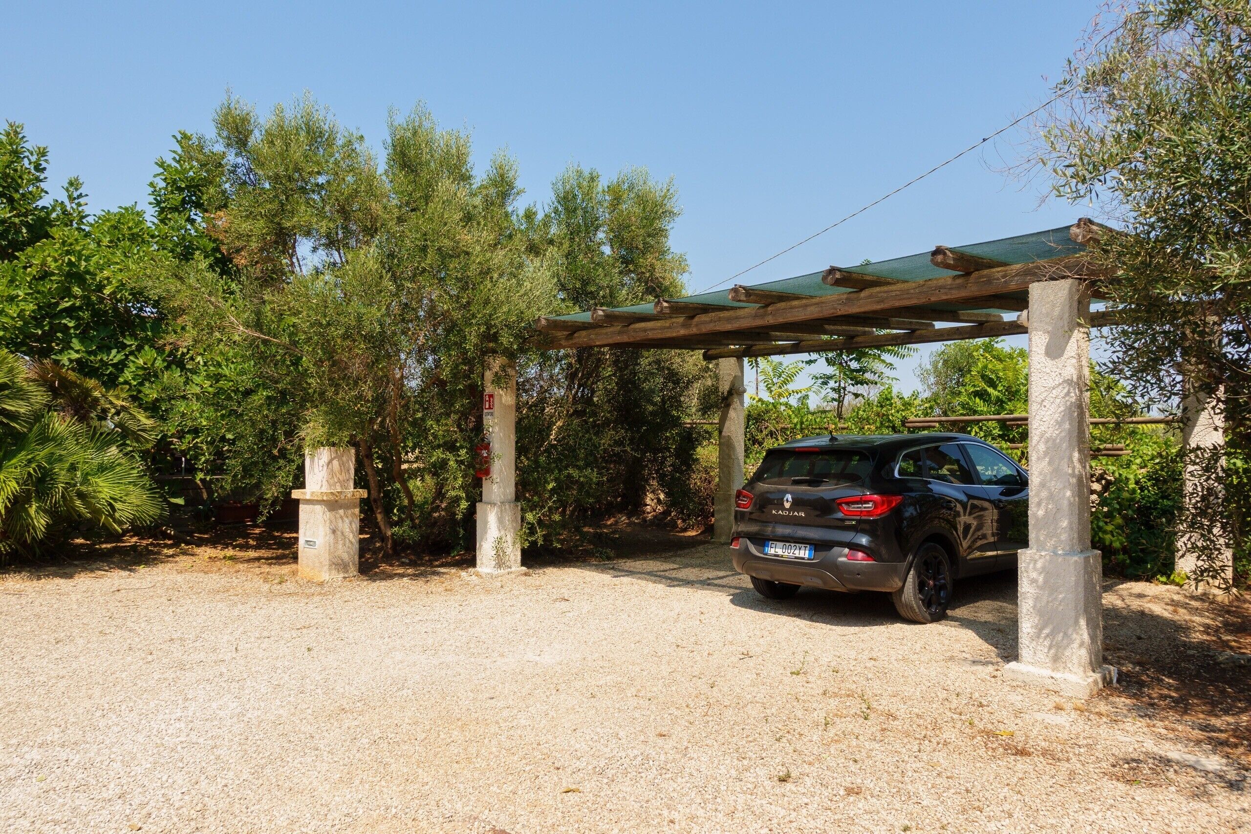 Une voiture est garée sous une pergola en bois, dans un espace recouvert de gravier et bordé d’arbres.