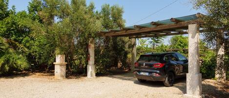 A car parked under a wooden pergola in a gravel area with trees.