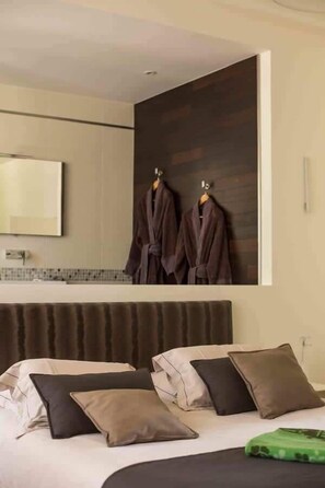 A modern hotel room with a large bed, multiple pillows, a headboard, a mirror, and two robes hanging on hooks.