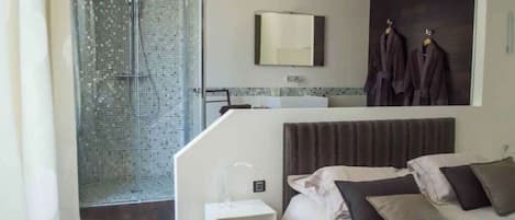 Een moderne hotelkamer met een douche, een bed met kussens, een klein nachtkastje en een spiegel boven het bed.