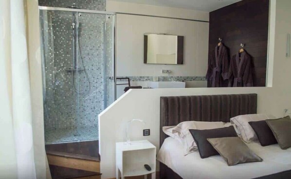 Una habitación de hotel moderna con ducha, una cama con almohadas, una mesita de noche pequeña y un espejo sobre la cama.