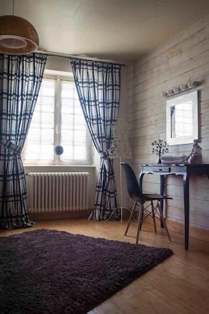 Une pièce avec un plancher en bois, un bureau avec une chaise, un radiateur et une fenêtre avec des rideaux.