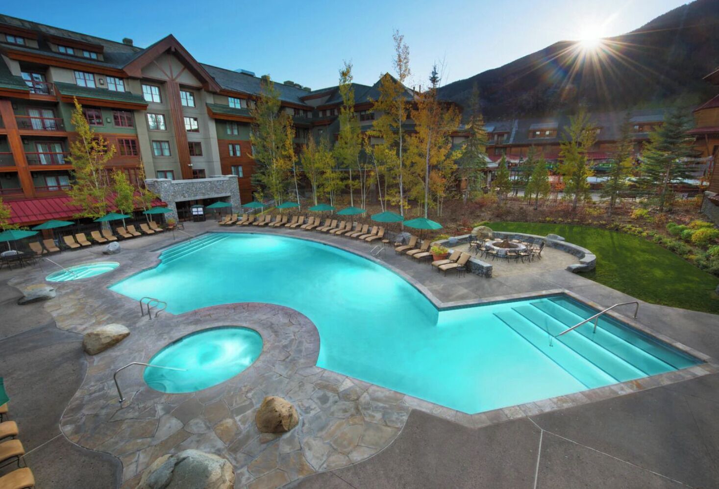 Une piscine de complexe hôtelier avec des chaises longues et un jacuzzi, entourée d’immeubles et d’arbres.