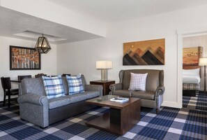 A modern living room with a grey sofa, a smaller sofa, a coffee table, and a checkered floor. There is a painting on the wall and a lamp on a side table.