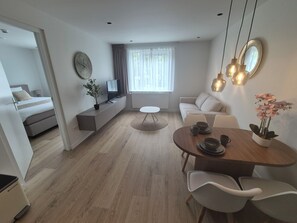 Ein modernes, helles Wohnzimmer mit einer Couch, einem Esstisch mit Stühlen, einem Fernseher auf einem Schrank und einer Topfpflanze.