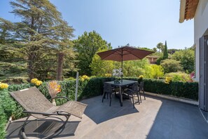 Un patio al aire libre con una mesa y sillas, un paraguas grande, y un jardín con plantas y flores de distintos tipos.