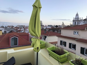 A balcony with a green umbrella, a potted plant, and a view of a cityscape with red-roofed buildings.