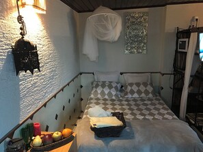 A room with a bed, a nightstand with a basket, a wall-mounted light fixture, and a decorative wall piece.