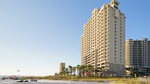 Un grand hôtel avec de nombreux balcons, situé près d’une plage où l’on peut voir des bateaux et un quai.
