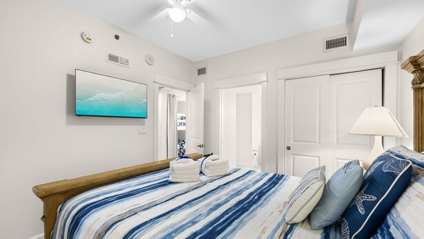 A bedroom with a bed, a ceiling fan, a mounted TV, and a door leading to a bathroom. - Ocean Breeze! 5th Floor 2BR Gulf-Front Condo Sleeps 6 (Panama City Beach)