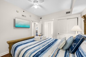 Ein Schlafzimmer mit Bett, Deckenventilator, an der Wand montiertem Fernseher und einer Tür zu einem Badezimmer.