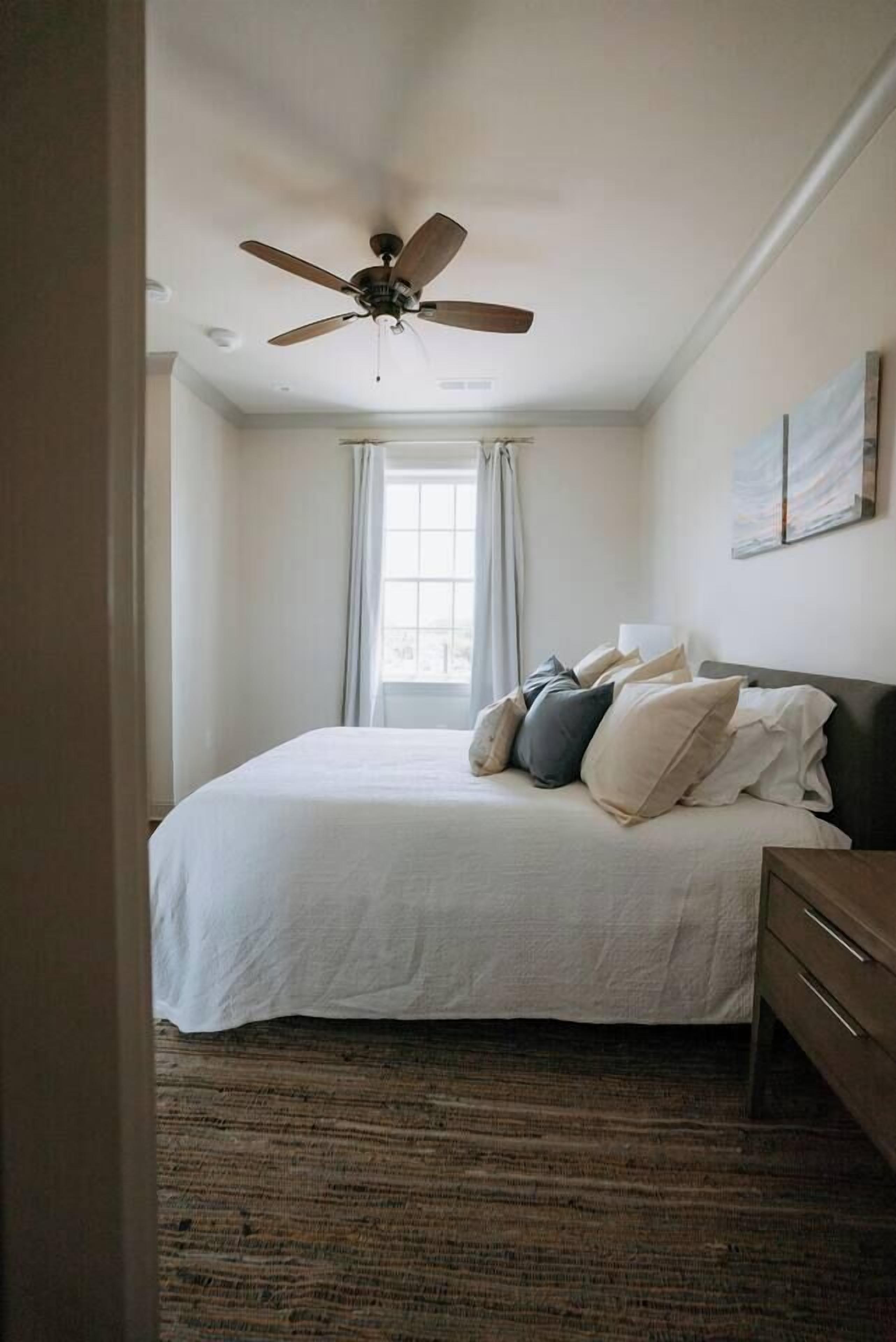 A bedroom with a bed, a ceiling fan, a window with curtains, and a wall-mounted artwork.