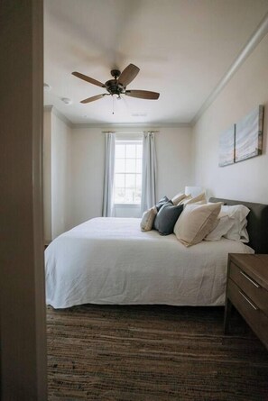 A bedroom with a bed, a ceiling fan, a window with curtains, and a wall-mounted artwork.