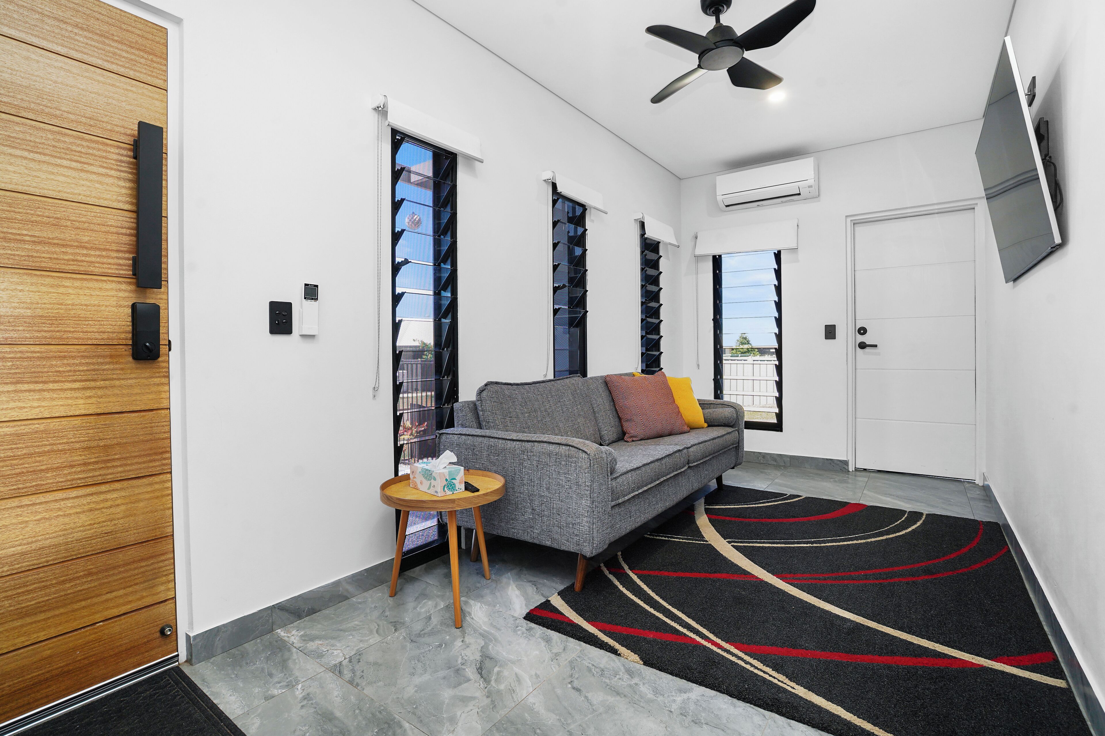 Ein modernes Interieur mit einem grauen Sofa, einem kleinen runden Tisch, einem Deckenventilator und einem an der Wand montierten Fernseher.